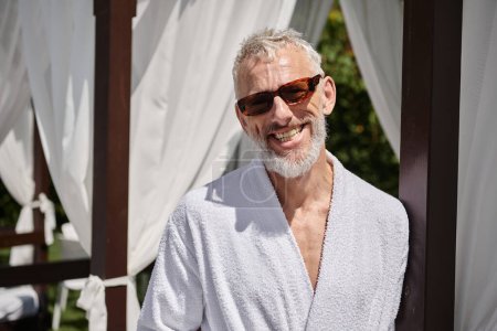 fröhlicher älterer Mann in Sonnenbrille und Mantel entspannt sich im privaten Pavillon, Rückzug im Resort