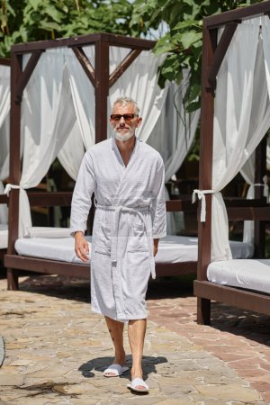 Mann mittleren Alters in stylischer Sonnenbrille und weißer Robe spaziert in der Nähe eines privaten Pavillons im Luxus-Resort
