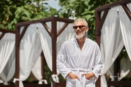 Téléchargez les photos : Joyeux homme d'âge moyen en lunettes de soleil et robe posant avec les mains dans les poches dans un complexe de luxe - en image libre de droit