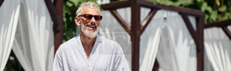 fröhlicher Mann mittleren Alters in stylischer Sonnenbrille sonnt sich im Luxusresort, horizontales Banner