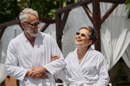 alegre pareja de mediana edad en gafas de sol y túnicas caminando en complejo de lujo, concepto de retiro de bienestar