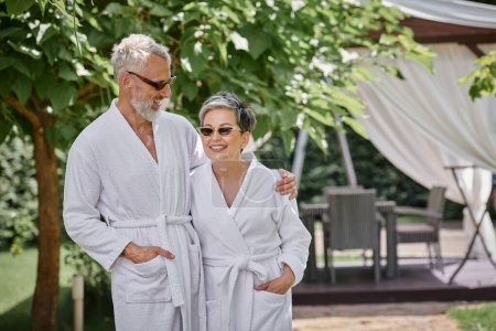 feliz pareja madura en gafas de sol y batas de pie en un resort de lujo, concepto de retiro de bienestar