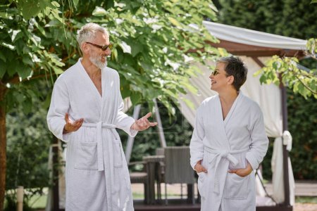 happy mature couple in sunglasses and robes chatting in luxury resort, wellness retreat concept