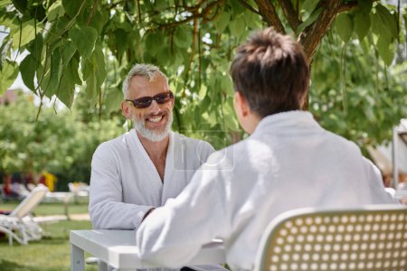 glücklicher reifer Mann in Sonnenbrille und Robe plaudert mit Frau im Sommergarten, Wellness-Rückzugsort