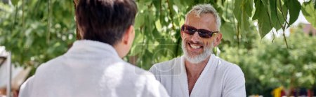 hombre maduro feliz en gafas de sol y bata charlando con la esposa en el jardín de verano, retiro, pancarta