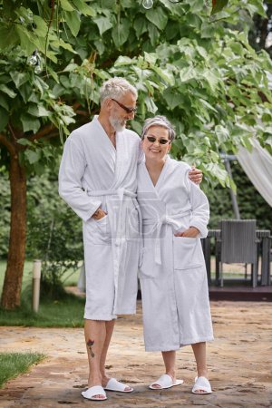 glücklicher reifer Mann mit tätowierter Ehefrau in Sonnenbrille und Robe im Sommergarten, Wellness-Rückzugsort