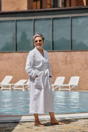 happy middle aged woman in white robe and sunglasses standing at poolside in luxury resort