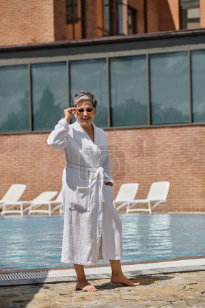 cheerful middle aged woman in white robe and sunglasses standing at poolside in luxury resort