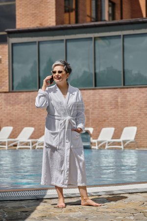 mujer de mediana edad feliz en bata y gafas de sol hablando en el teléfono inteligente junto a la piscina en el complejo de lujo