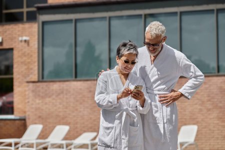 happy middle aged woman in sunglasses using smartphone near husband at poolside in luxury resort