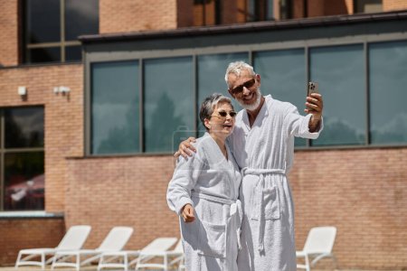 happy middle aged couple in robes taking selfie on smartphone in luxury resort, wellness retreat