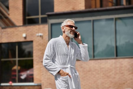 happy and tattooed middle aged man in sunglasses and robe talking on smartphone in resort, retreat