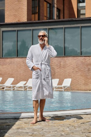 glücklicher und tätowierter Mann mittleren Alters in Sonnenbrille und Robe, der am Pool mit dem Smartphone spricht, sich zurückzieht