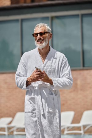 Foto de Hombre de mediana edad feliz y tatuado en gafas de sol y bata con teléfono inteligente en el resort, retiro - Imagen libre de derechos