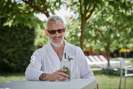 Wellness-Rückzugskonzept, glücklicher Mann mittleren Alters in Sonnenbrille und Bademantel genießt Cocktail im Urlaub