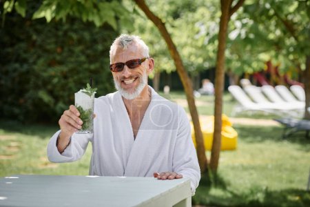 concept of wellness retreat, happy mature man in sunglasses and robe enjoying refreshing cocktail