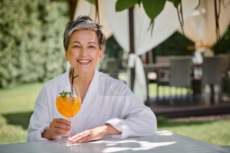 concept of wellness retreat, happy mature woman in white robe enjoying cocktail on vacation