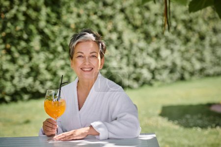 concepto de retiro de bienestar, alegre mujer de mediana edad en bata disfrutando de un cóctel de vacaciones