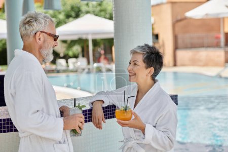 happy middle aged couple in sunglasses and robes holding cocktails at poolside, wellness retreat
