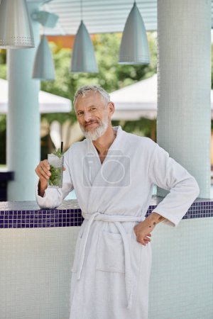 positiver reifer Mann in weißer Robe mit erfrischendem Mojito-Cocktail am Pool, Rückzug