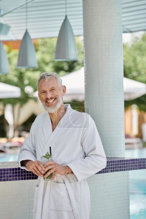 Mann mittleren Alters in weißer Robe lächelt und hält Mojito-Cocktail am Pool, Wellness-Rückzugsort