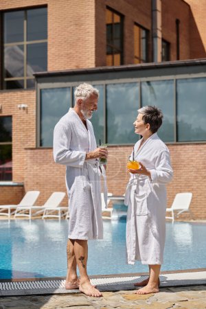 cheerful mature couple in white robes holding cocktails at poolside, wellness retreat in resort