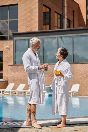 cheerful middle aged couple in white robes holding cocktails at poolside, wellness retreat in resort