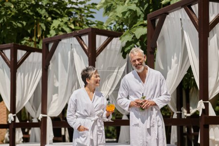 cheerful middle aged couple in white robes holding cocktails near pavilion, wellness retreat