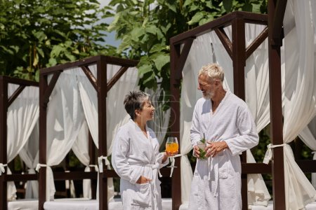 happy mature couple in white robes holding cocktails and talking near pavilion, wellness retreat