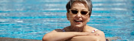 alegre mujer de mediana edad en gafas de sol y traje de baño dentro de la piscina, retiro, pancarta
