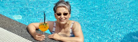 mujer madura feliz en gafas de sol nadando en la piscina con agua azul y la celebración de cóctel, pancarta