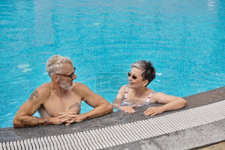 middle aged couple in sunglasses chatting inside of swimming pool during vacation, wellness retreat