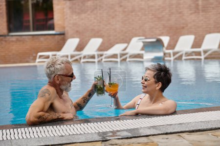 happy mature couple in sunglasses clinking cocktails inside of swimming pool, wellness retreat