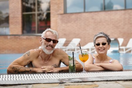 happy mature couple in sunglasses swimming in pool smiling near cocktails, wellness retreat concept
