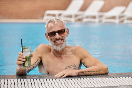hemdlos und tätowiert Mann mittleren Alters mit Sonnenbrille hält Cocktail und schwimmt im Pool, Rückzug
