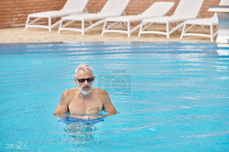 shirtless and tattooed middle aged man in sunglasses swimming in pool with blue water, retreat