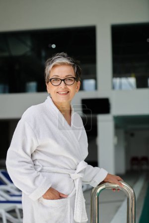 happy middle aged woman in white robe and glasses smiling inside of indoor spa center, retreat