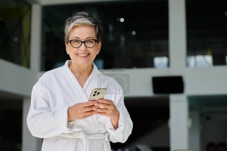 glückliche reife Frau in weißer Robe und Brille mit Smartphone im Indoor-Wellness-Center, Rückzug