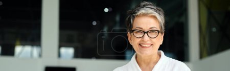 happy mature woman in white robe and glasses looking at camera inside of indoor spa center, banner