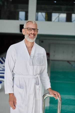 happy mature man in white robe and glasses smiling inside of indoor spa center, wellness retreat