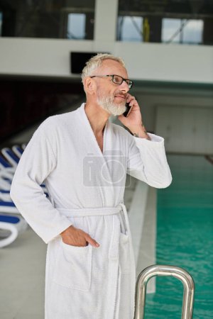 tattooed mature man in white robe and glasses talking on smartphone inside of spa center, retreat