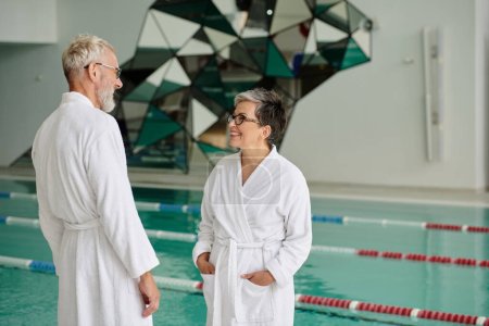 Photo for Cheerful middle aged couple in glasses and white robes chatting near indoor pool in spa center - Royalty Free Image