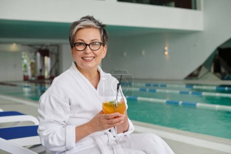 spa center concept, happy mature woman in glasses and robe holding cocktail and sitting on lounger