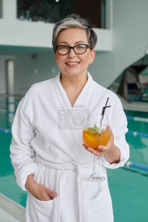 spa and retreat concept, happy mature woman in glasses and robe holding cocktail near indoor pool