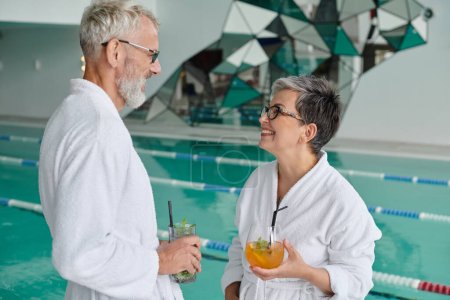 cheerful middle aged couple in white robes holding glasses with cocktails in spa center near pool