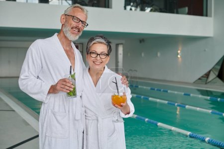 happy mature couple in white robes holding glasses with cocktails in spa center near indoor pool