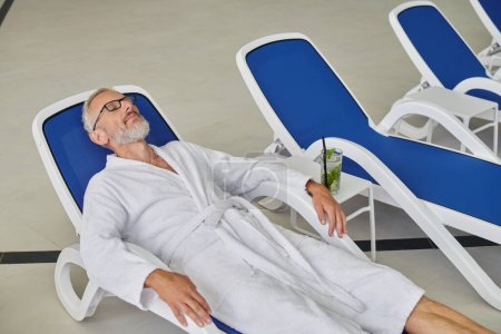 Wellness-Konzept, reifer Mann in Brille und Mantel ruht auf Liege in der Nähe von Cocktail im Kurzentrum