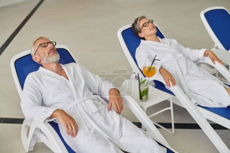 concepto de retiro, pareja madura en túnicas blancas descansando en tumbonas cerca de cócteles en el centro de spa