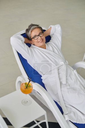 retreat concept, happy mature woman in white robe resting on lounger near cocktail in spa center