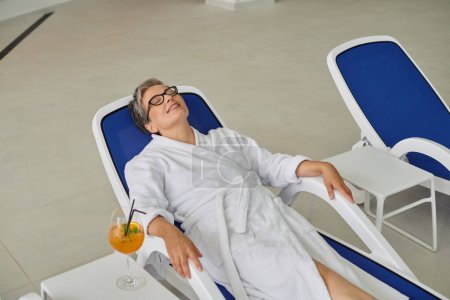 Rückzugskonzept, erfreut reife Frau in weißem Gewand auf Liegen in der Nähe von Cocktail im Wellness-Center ausruhen
